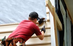 Young homeowner installs siding to his home. He is holding a hammer and wearing a tool belt.