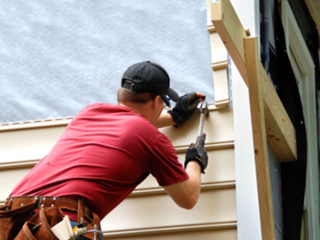 Young homeowner installs siding to his home.  He is holding a hammer and wearing a tool belt.
